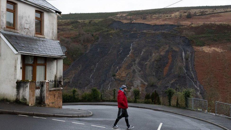 Man walks past house in Tylorstown, with coal tip landslide visible behind on hillside