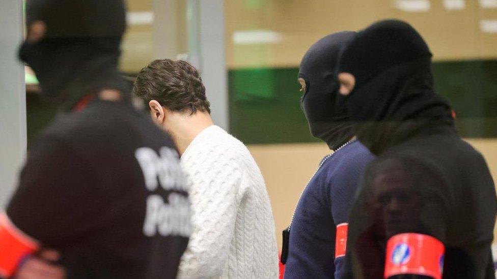 A man in a white jumper is surrounded by policemen wearing balaclavas