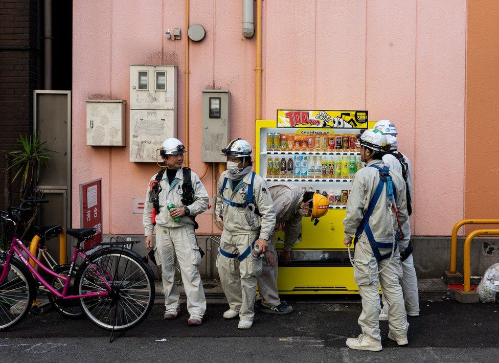 workmen with helmets and body suits grab a drink from a vending machine