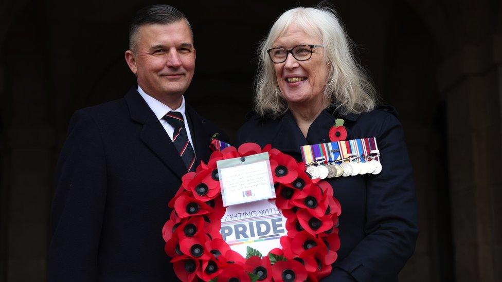 A man and woman with military medals hold a poppy wreath that says 'Fighting With Pride.'