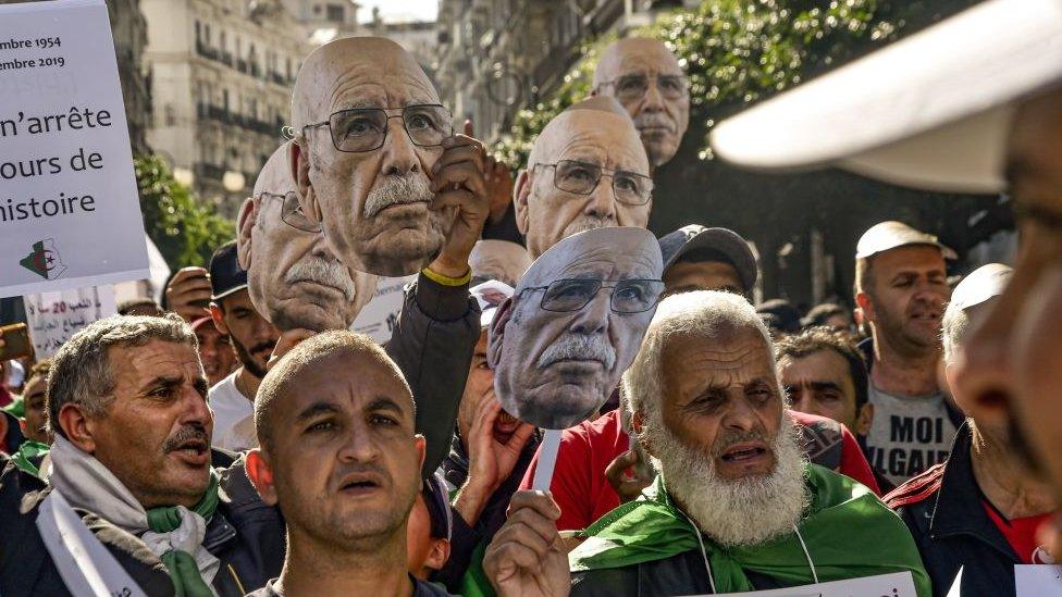 Protesters holding masks showing the face of Lakhdar Bouregaa