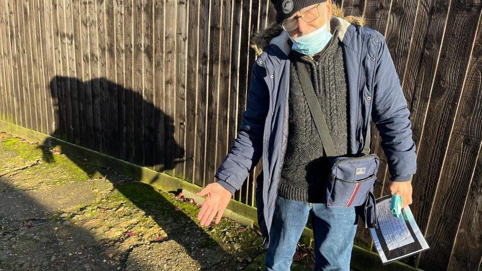 Man wearing blue jacket, mask and woollen hat points to cracks in alleyway surface