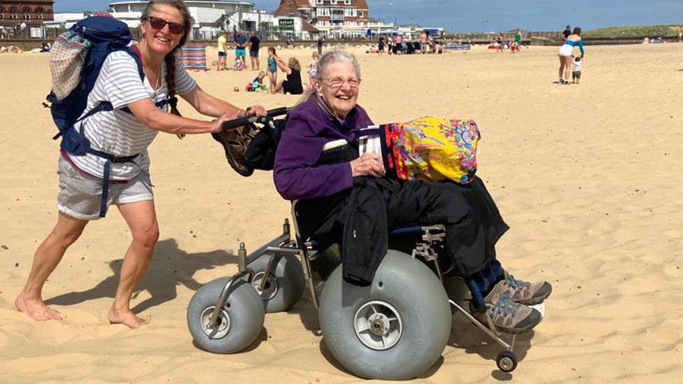 Woman pushing an older woman in a beach wheelchair with large grey pneumatic tyres on a sandy beach