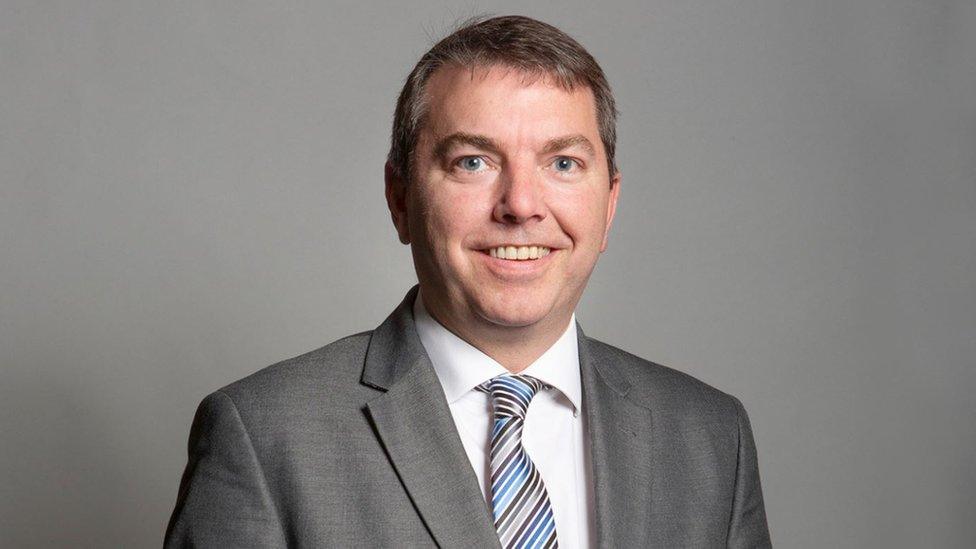 A middle-aged white man with short dark hair wears a grey suit with a white shirt and blue tie and smiles at the camera