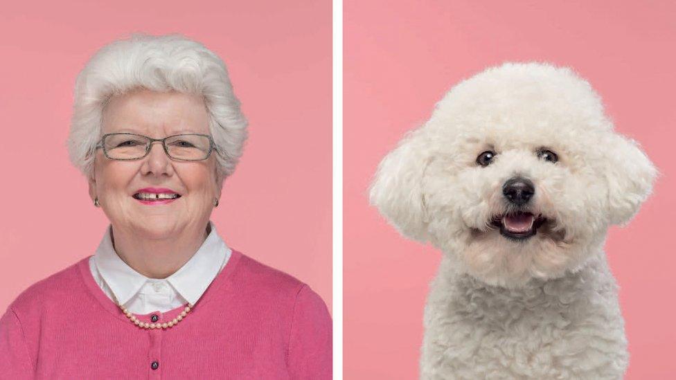 A woman with a bright smile and grey glasses looks forward next to a fluffy white dog which looks like its smiling