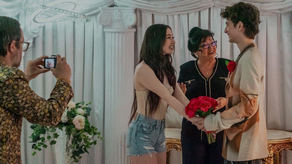 Mikey Madison and Mark Eidelstein in Anora. Their characters appear to be getting married - although it seems like a Las Vegas style wedding. They are looking at one another and holding a bouquet of roses while the woman officiating the ceremony looks at them and smiles. 