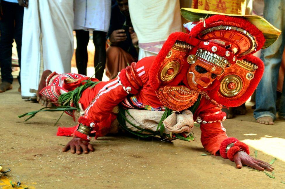 A performer lays stomach-down on the floor. They are wearing a red headdress and costume