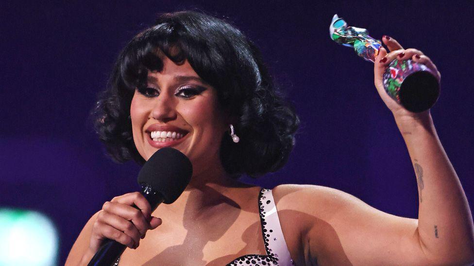 A smiling Raye holding up a Brit Award on stage in 2024