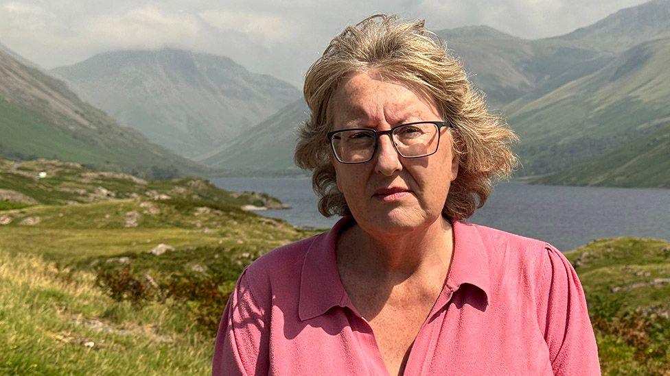 Jane Hall is standing half way up Scafell Pike in Cumbria. Behind her are the fells and Wast Water. She is wearing a pink polo shirt and has glasses. Her grey-blonde chin-length hair is being blown back off her face by the wind. She looks pensive.