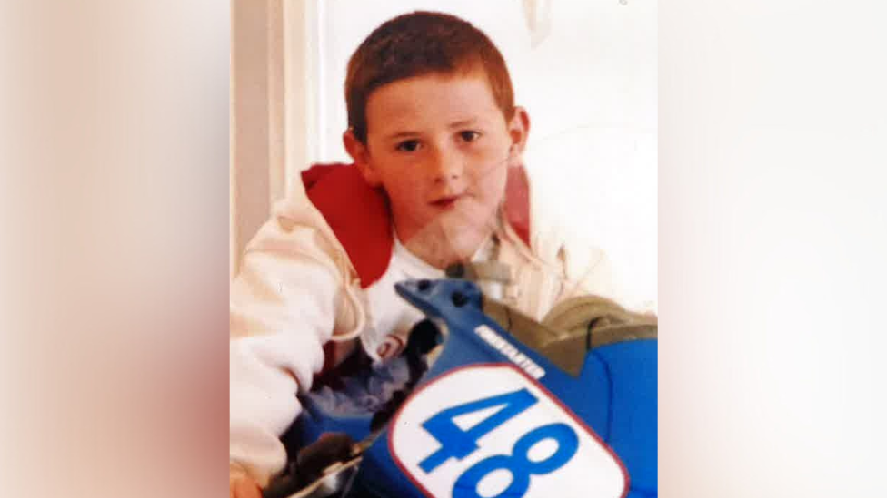 A photograph of Anthony Binfield as a child, wearing a white hoody with a red lining on a toy motorbike with the number '48' on the front.