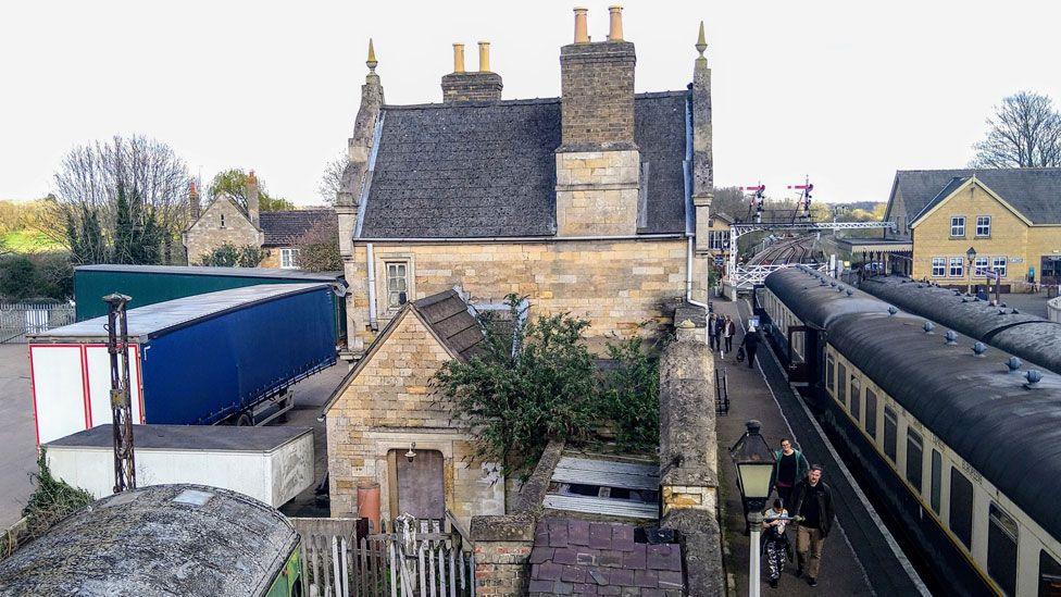 Wansford Station, part of the Nene Valley Railway. The two-storey building is shown, built from cream coloured brick and with a peaked grey tiled roof. The platforms are on the right, with carriages waiting on both sides.