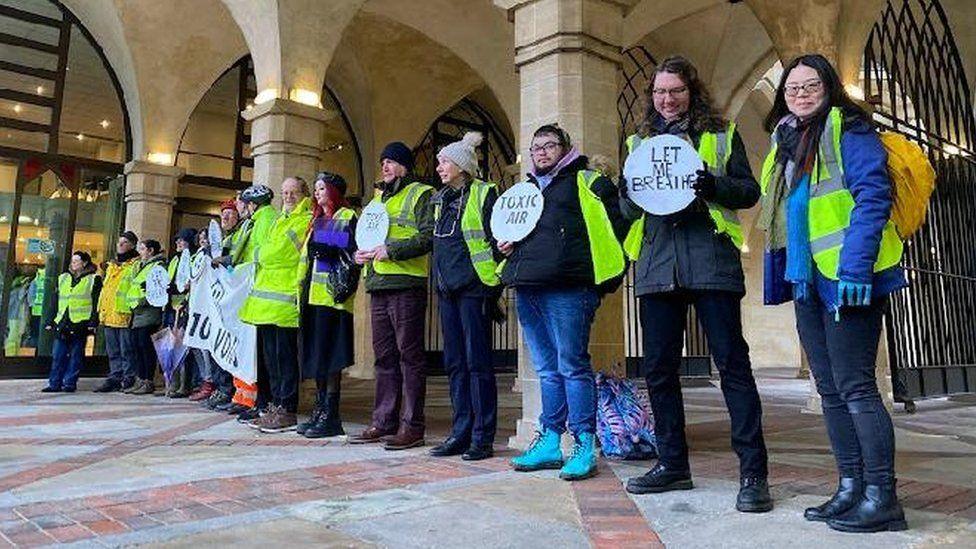 A line of people, some wearing yellow hi-vis vests, and many carrying circular placards with air quality slogans. They are in front of the stone pillars of the exterior of Northampton Guildhall