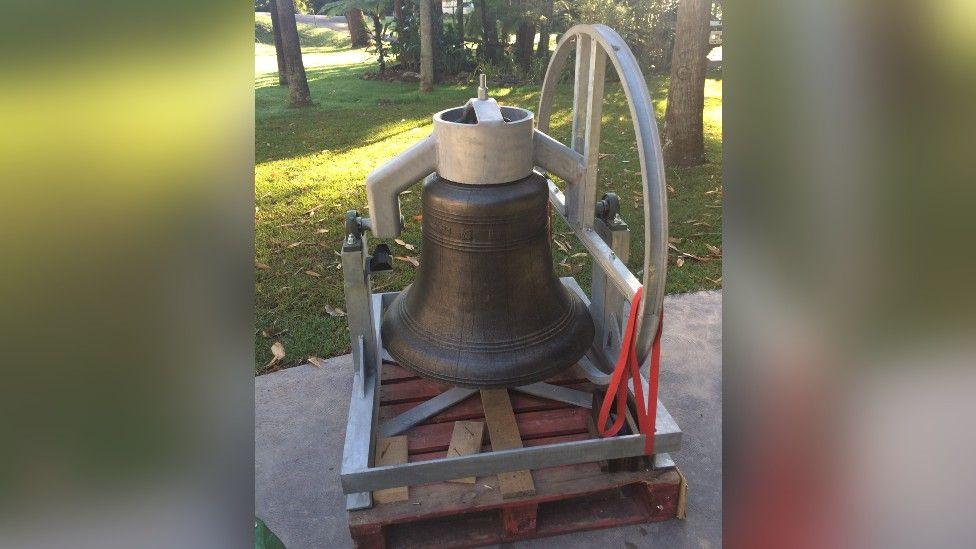 The bell in Australia before it was installed in the tower. It is on a driveway with grass and trees in the background.