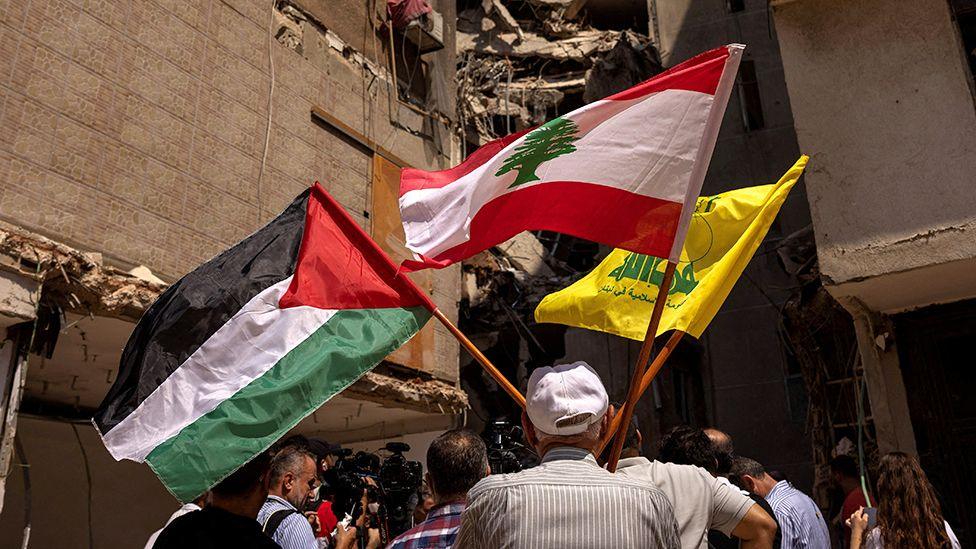 A man holds Palestinian, Lebanese and Hezbollah flags next to a damaged site where top Hezbollah commander Fuad Shukr was killed in Beirut in August