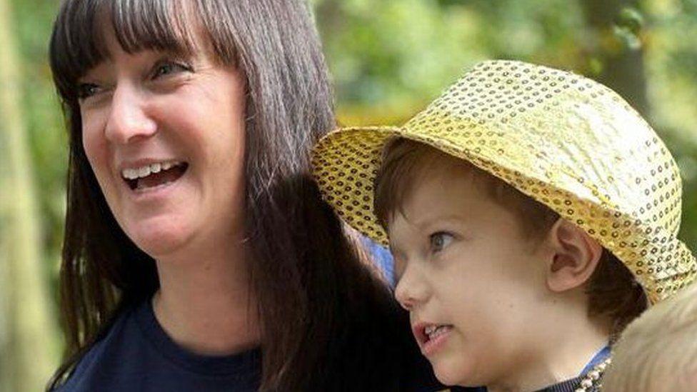 Lisa Radcliffe with shoulder length black hair smiling while holding George, who is wearing a large yellow hat 