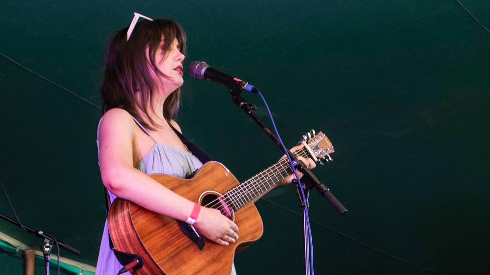 A woman in a purple dress sings into a microphone while playing guitar