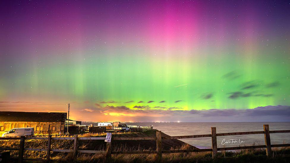 The Northern Lights over the sea at Mappleton in East Yorkshire with buildings and a fence in the foreground