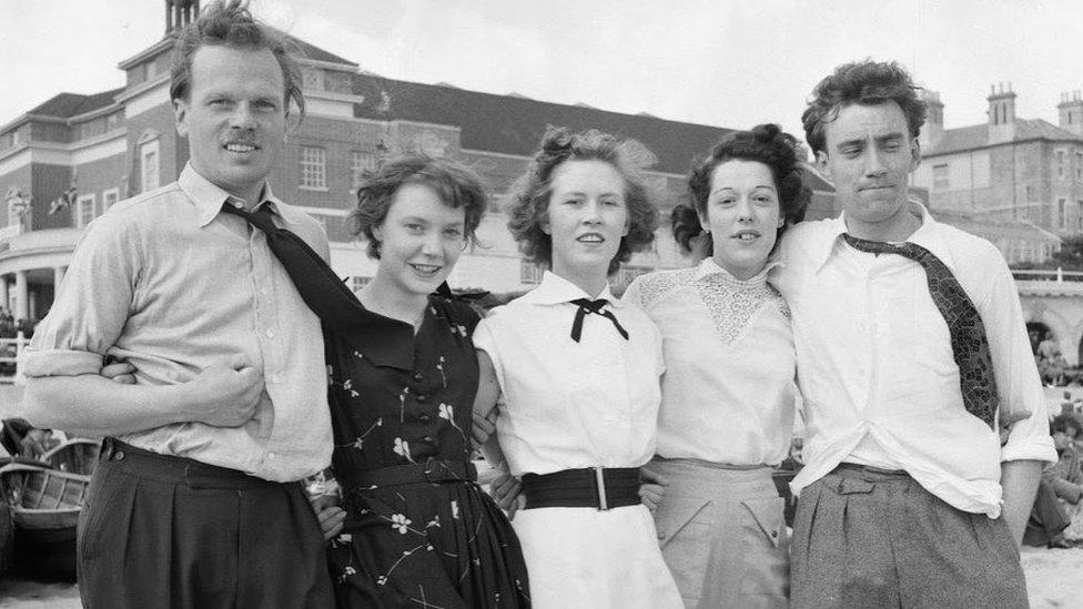 Two men and three women link arms on the beach