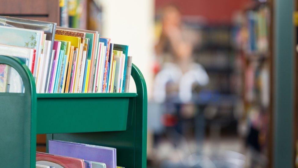 Green rolling cart full of colourful children's books sitting in front of large bookshelves in modern public library or elementary school library.