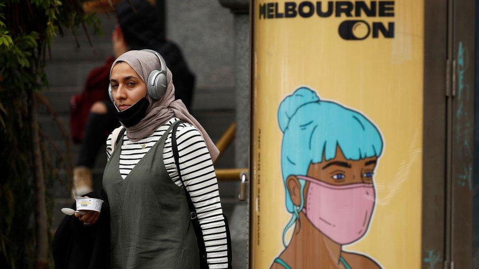 A young woman wearing a mask and a hijab walks past a pole with an ad featuring a graphic illustration of a woman with blue hair wearing a mask