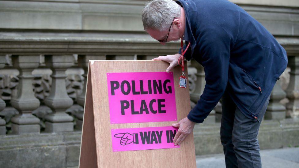 A man with grey hair, wearing dark clothes, bends over a sign that says "polling place". The sign is on the pavement of a street, with a stone railing in the background.