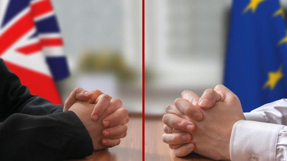 Two men's hands across a negotiating table, in front of UK and EU flags