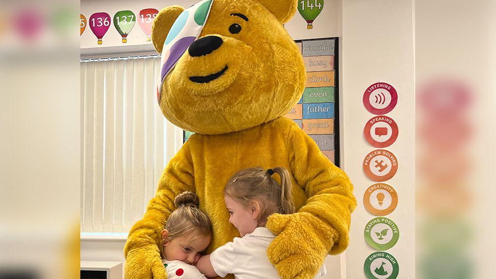 Two children in spotty T-Shirts hugging Pudsey