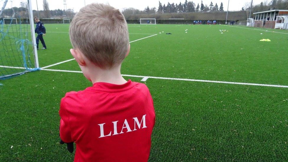 Liam facing away from the camera looking out at a pitch. On the back of his red shirt is his name in capital letters, akin to the names on the back of footballers' shirts.