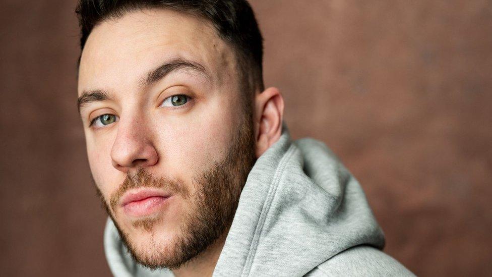 Louis Stannett is a 26-year-old white man with short brown hair and a short beard. He has green eyes and looks at the camera. He wears a grey hoody and is pictured against a brown backdrop