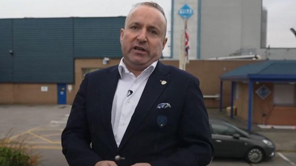 Dave Fishwick standing outside a factory with blue cladding and brinde brick walls. He is wearing a white shirt and dark blue suit.