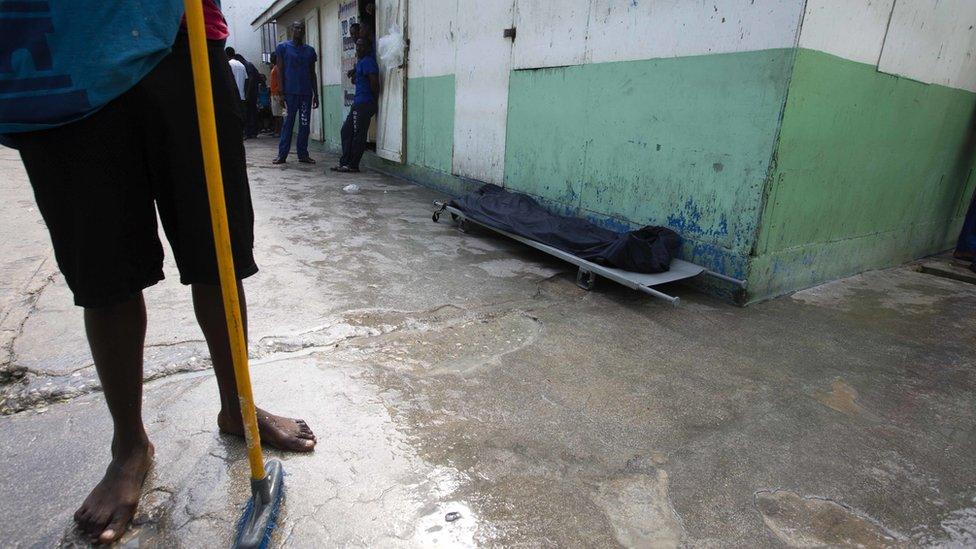 A prisoner stands near the body of an inmate, covered with a plastic tarp, who died of malnutrition inside the National Penitentiary in downtown Port-au-Prince