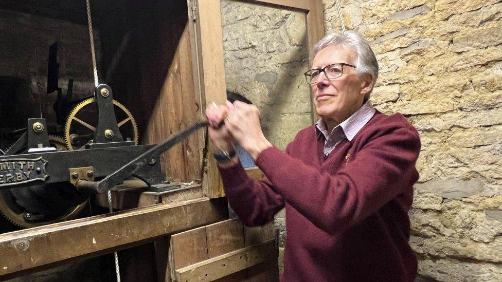 Man wearing red sweater pulls on the handle of a clock mechanism inside a stone tower