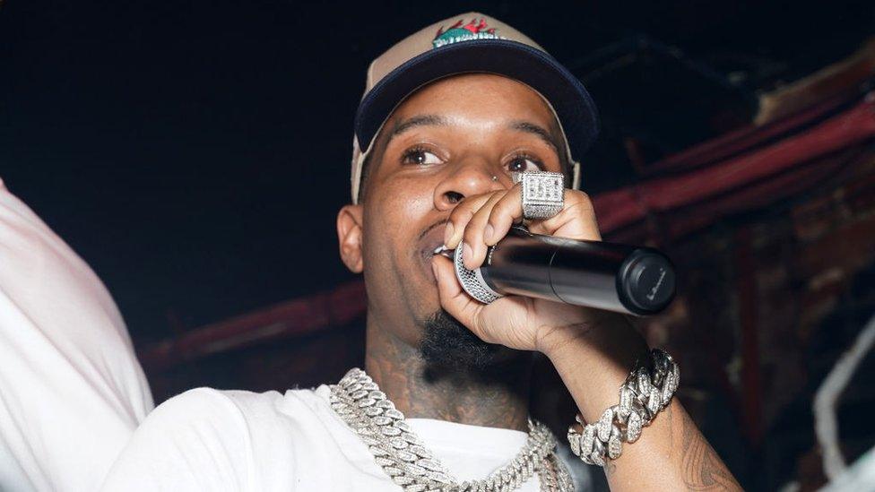 Tory Lanez rapping at the Sorry For What Event on September 28, 2022 in New York City. He's wearing a chunky, expensive-looking silver bracelt and matching necklace. He's holding a microphone close to his face, and the side of a very large, diamond-studded ring on his pinky finger is visible.