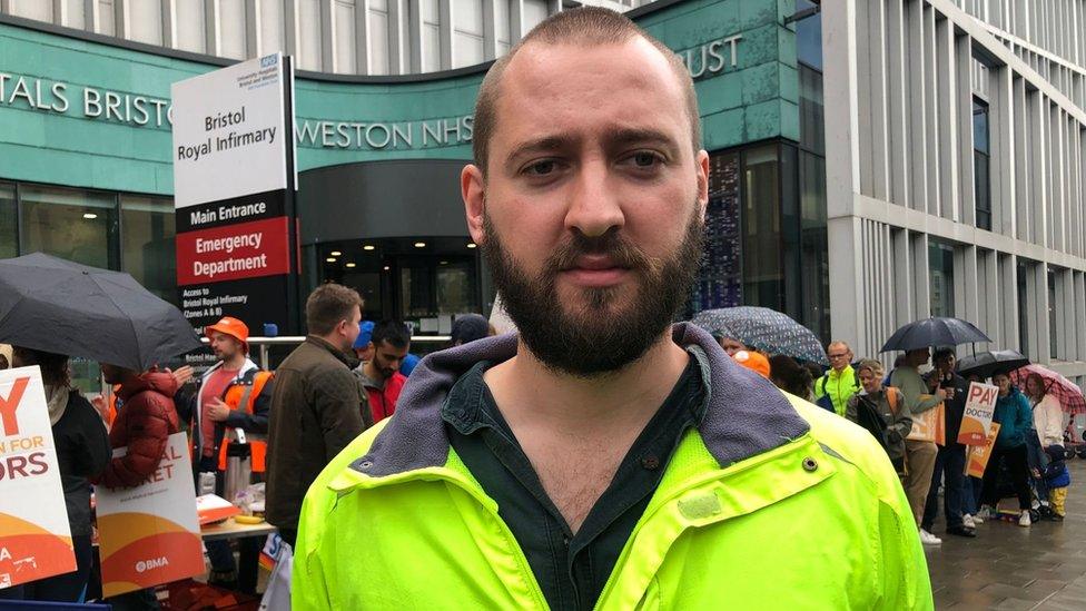 Dr Sam Taylor-Smith standing in front of the BRI with people striking in the background