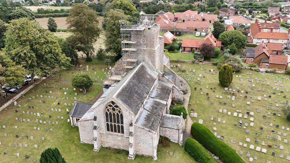 Drone image of St Mary's Church in Heacham showing scaffolding around the tower, with the cupola visible on top, and gravestones in the churchyard. Modern housing is in the background.