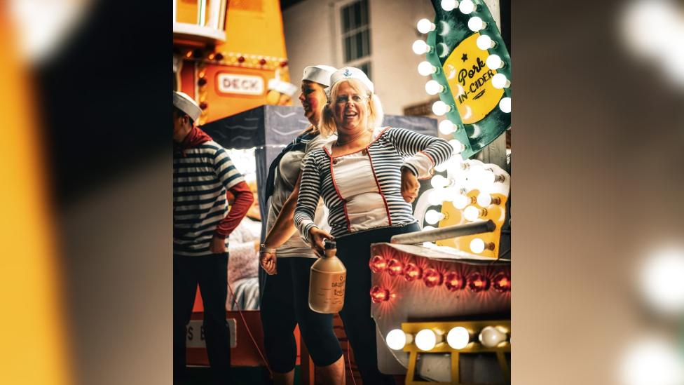 A woman wearing a costume standing on one of the carnival floats and smiling, holding a jug of beer