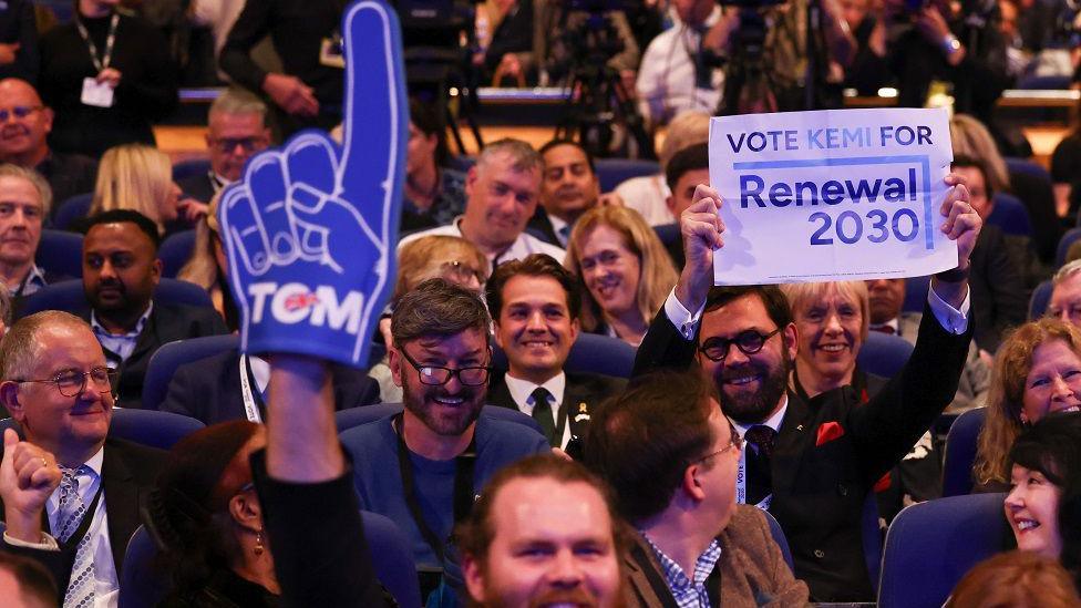 Bearded man in the audience at the Conservative Party conference waves Tom Tugendhat foam finger