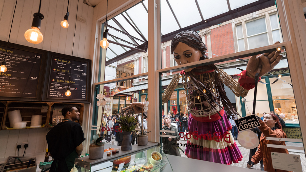 Little Amal peaks through a bakery window to scope out a bite to eat