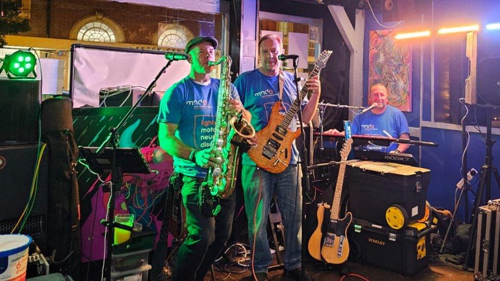 Mark and Ian wearing blue 'fighting motor neurone disease' shirts whilst performing on stage. One is playing the saxophone and the other is playing a guitar.