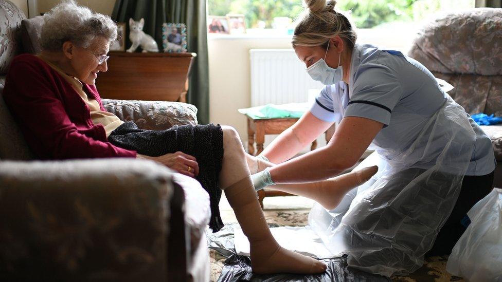 a nurse Rebecca, wearing personal protective equipment (PPE), changes the dressings on the legs of an elderly woman during a home visit.