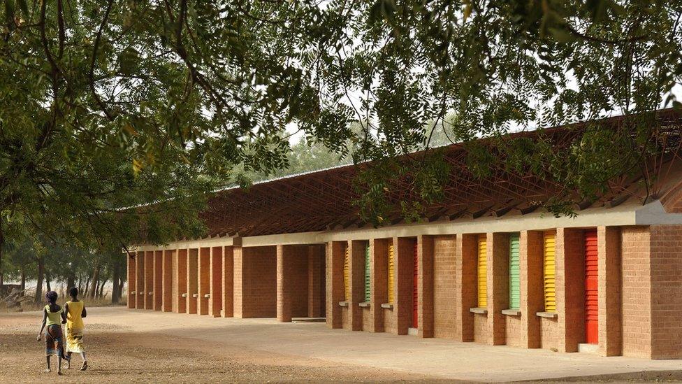 Gando Primary School. There are colourful shutters and trees in the background. There are two girls walking past it.