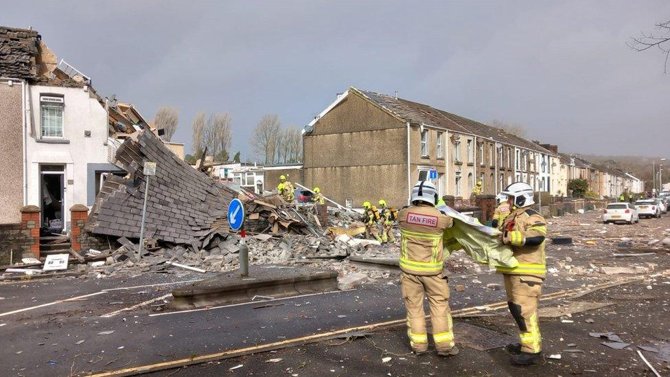 Emergency workers are stood amidst the rubble of the collapsed house. The upper floor of the house is exposed and leaning down to the ground while officers remove rubble.