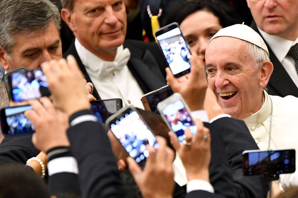 Pope Francis smiles as people take pictures on their mobile phones