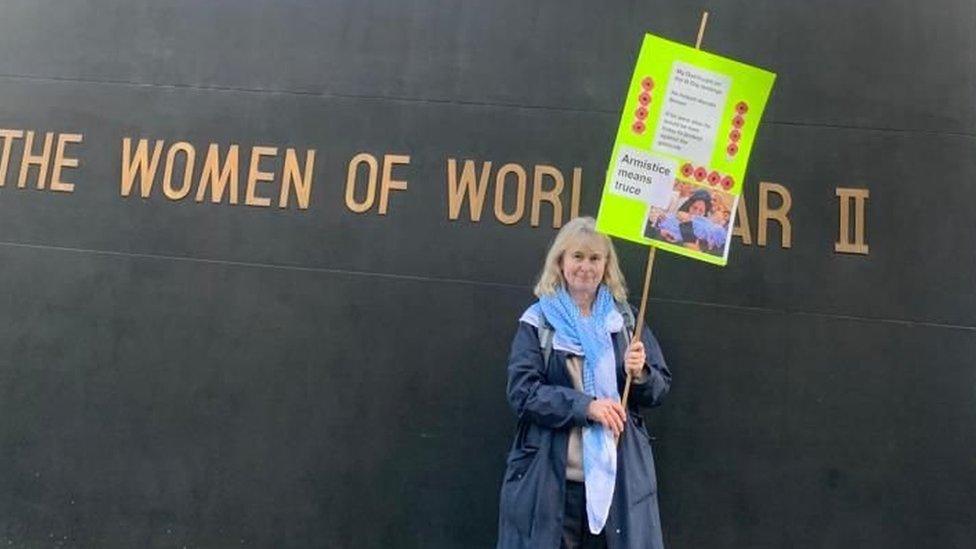 Ms Webster holding a yellow placard reading 'Armistice means truce' in front of a World War 2 memorial