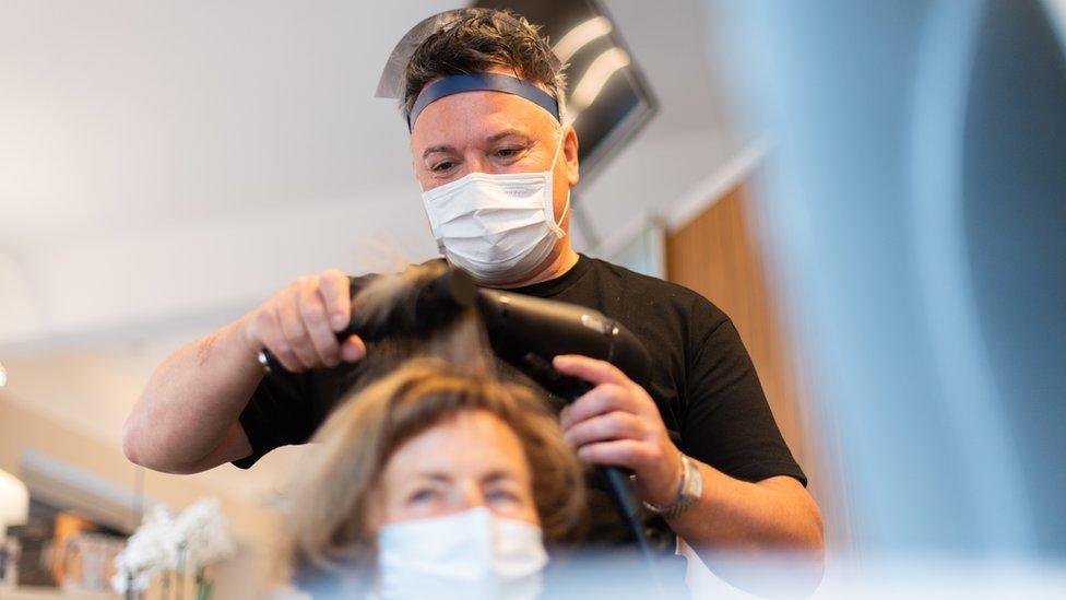 Hairdresser does the hair of a customer in his hairdressing studio, wearing a mouth guard and face shield.