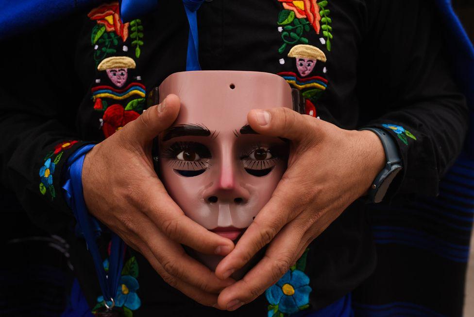 A close-up view  of a persons hands holding the traditional mask of Parachico - a man's face with black eye make-up.