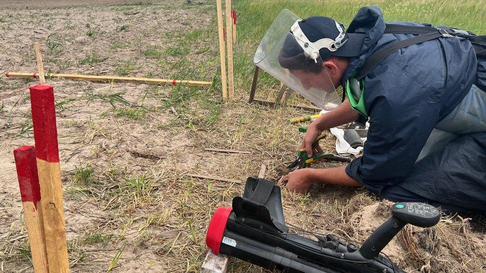 A HALO Trust worker on the ground wearing a helmet and vest using equipment.