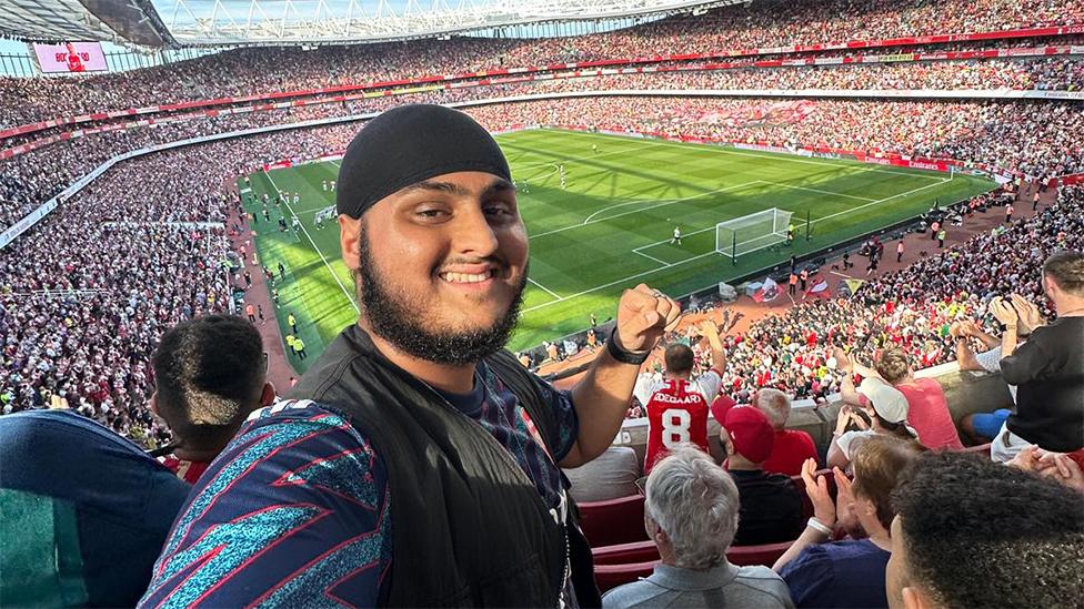 Bhavs, a South Asian man at Arsenal's Emirates stadium. He is wearing a black vest top with a light and dark blue striped top underneath. His left hand is raised in a fist pump action. Behind him is a green football pitch with thousands of fans in the stadium around him.