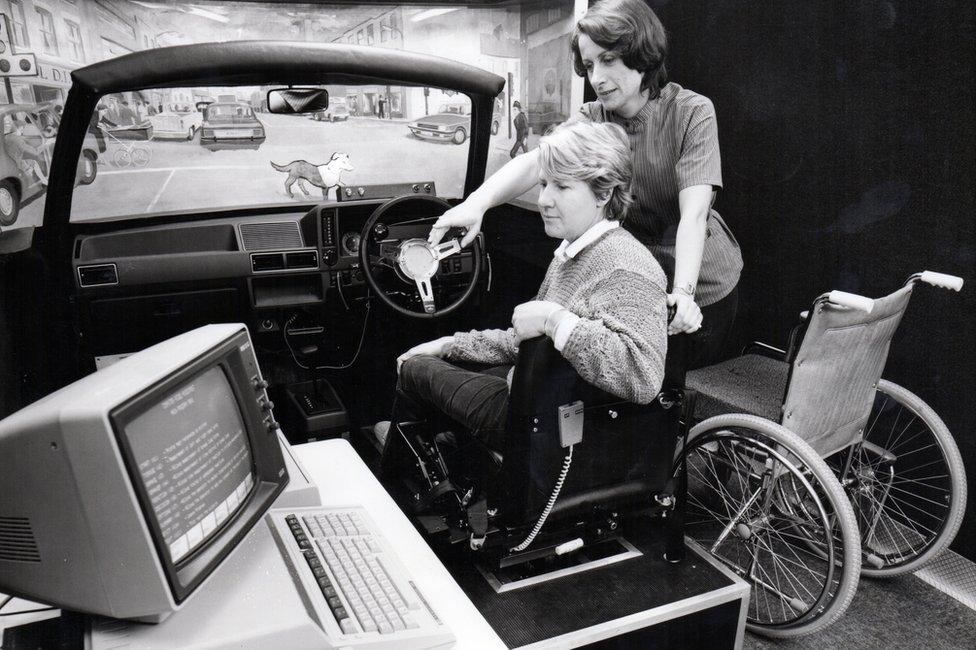 A woman demonstrating how an adapted car works. They are looking at an old computer screen