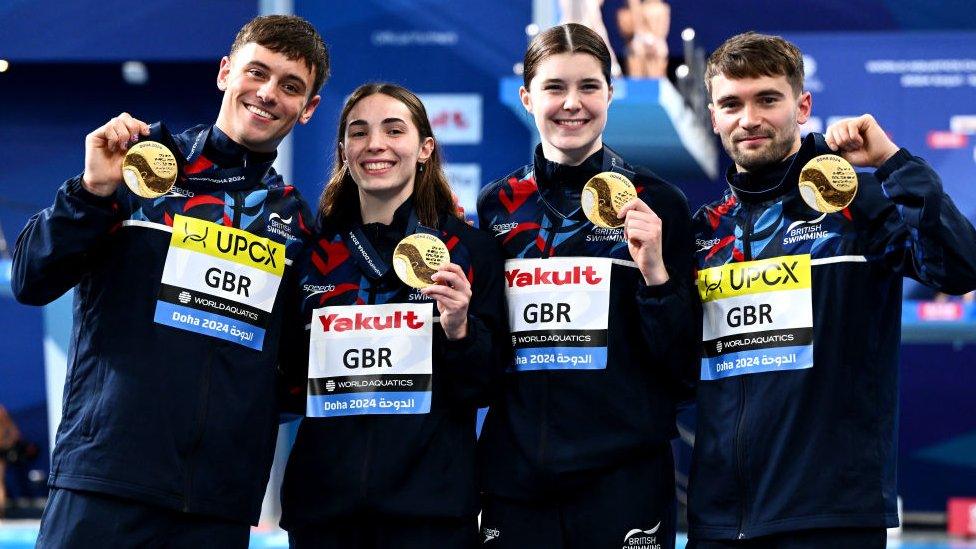 Gold Medalists, Daniel Goodfellow, Thomas Daley, Scarlett Mew Jensen and Andrea Spendolini Sirieix of Team Great Britain pose with their medals during the Medal Ceremony after the Mixed Team Event Final on day one of the Doha 2024 World Aquatics Championships. The team all wear navy blue British Swimming tracksuits and smile at the camera.